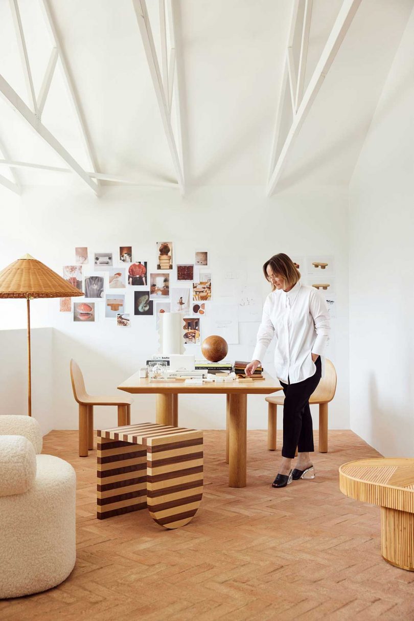 Sarah Ellison dressed in black and white stands at her wood desk in vaulted white studio