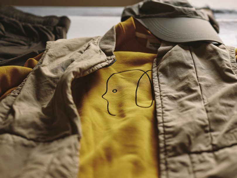 Detail of Norse Projects x McFetridge khaki vest over yellow sweatshirt, topped with a cap.