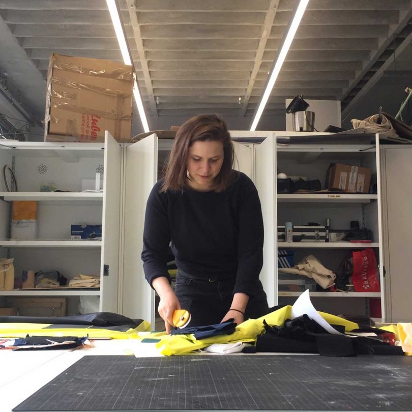 woman with dark hair and black shirt on standing leaning over work table cutting materials