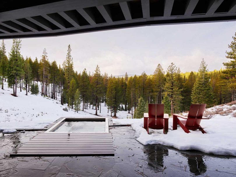 cover patio of modern house looking out past hot tub and two red chairs to snowy landscape