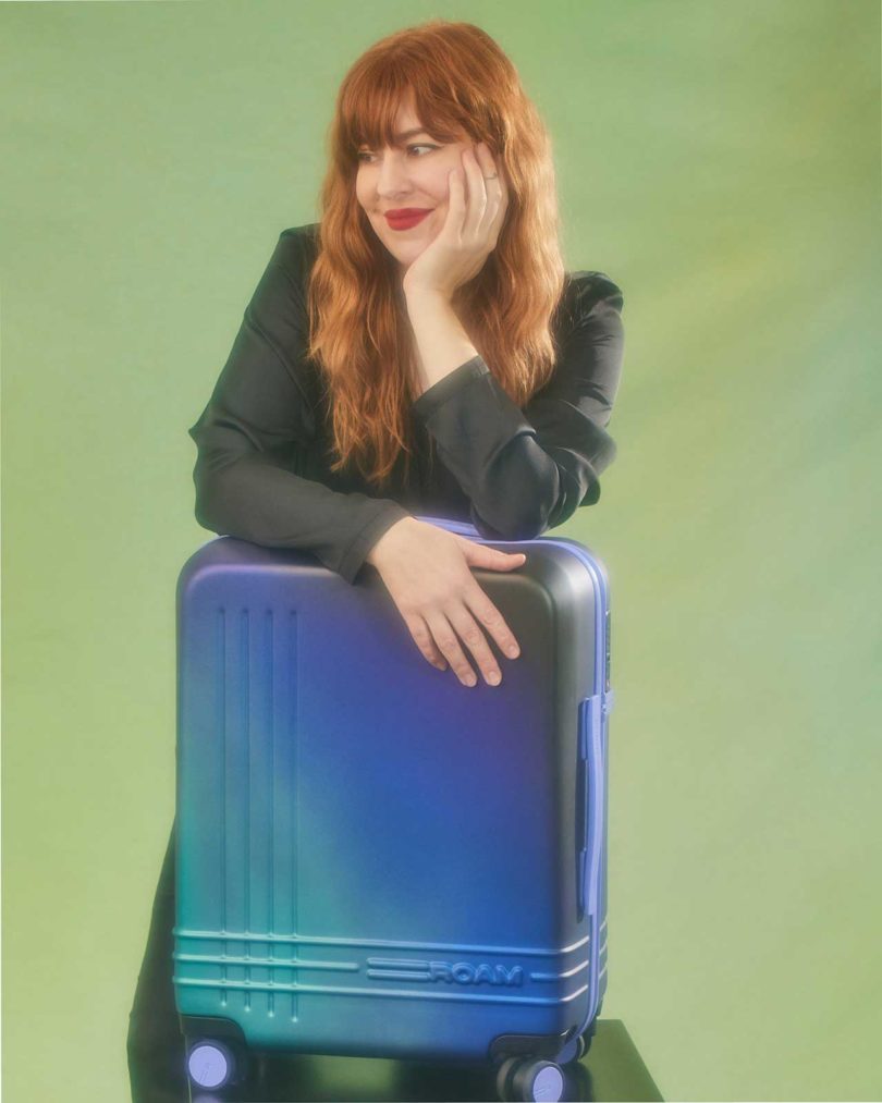 red-headed tattoo artist Amanda Wachob leaning on hand-painted iridescent suitcase