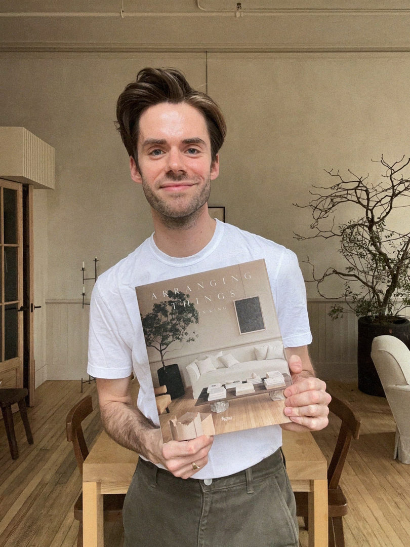 light-skinned man with dark hair wearing a white t-shirt and holding out a book in front of him