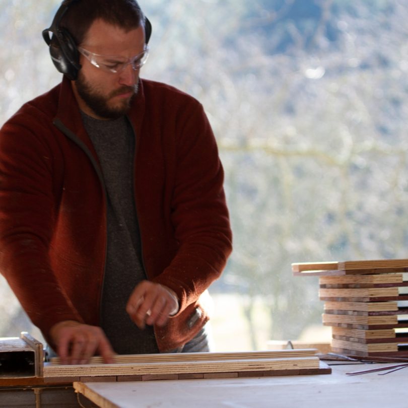 A white man with eye protectors and ear defenders is sanding a plank of wood. A pile of sanded planks is to his left. 