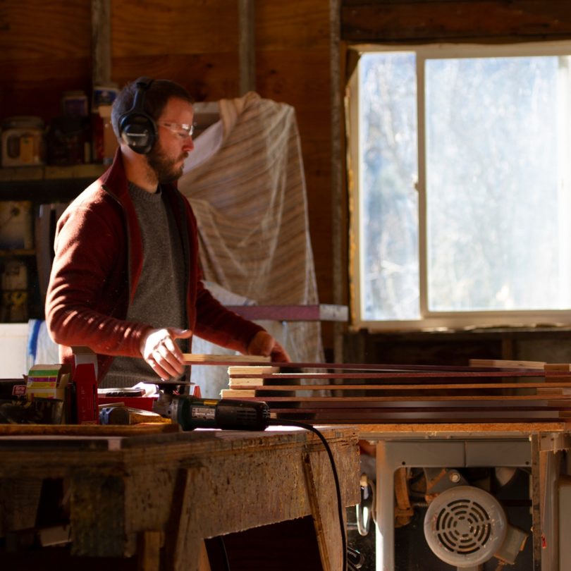 A white man with eye protectors and ear defenders sands wood in a workshop next to a window.