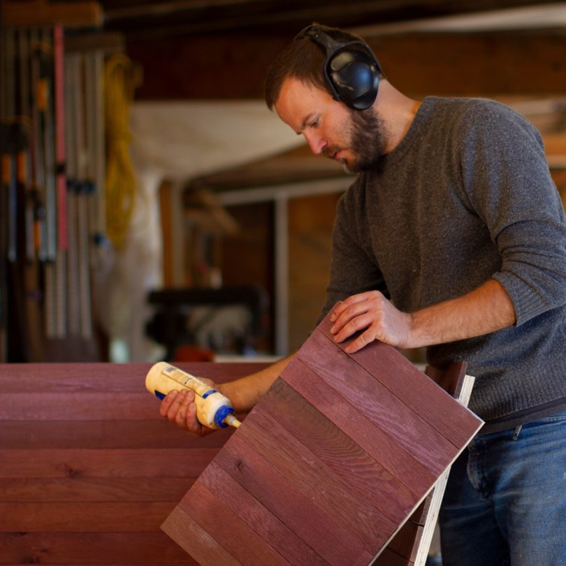 A white man with a beard and ear defenders glues the side of a piece of wood.