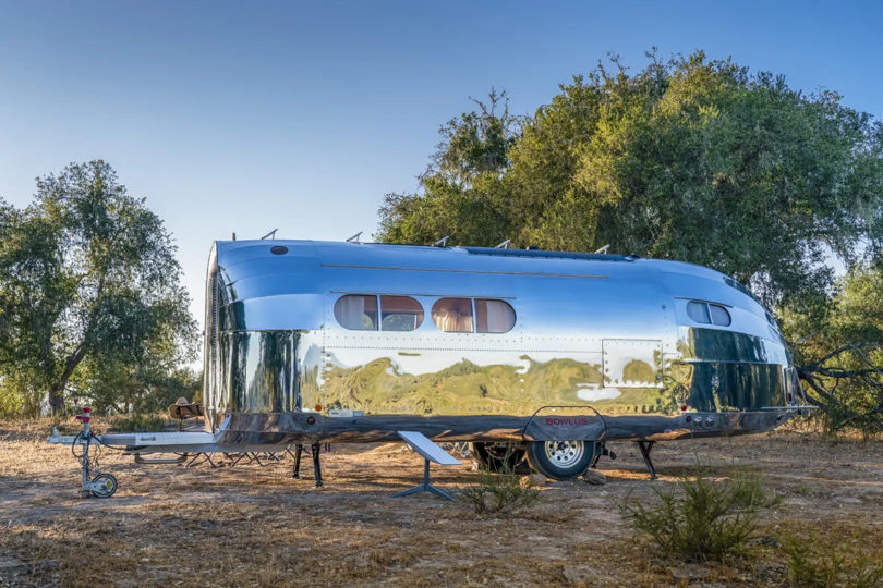 Side profile of Bowlus Volterra parked in campground during daytime under blue sky and surrounded by trees.