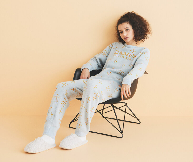 girl sitting slouched in black chair in pale grey lounge suit with eames written on it