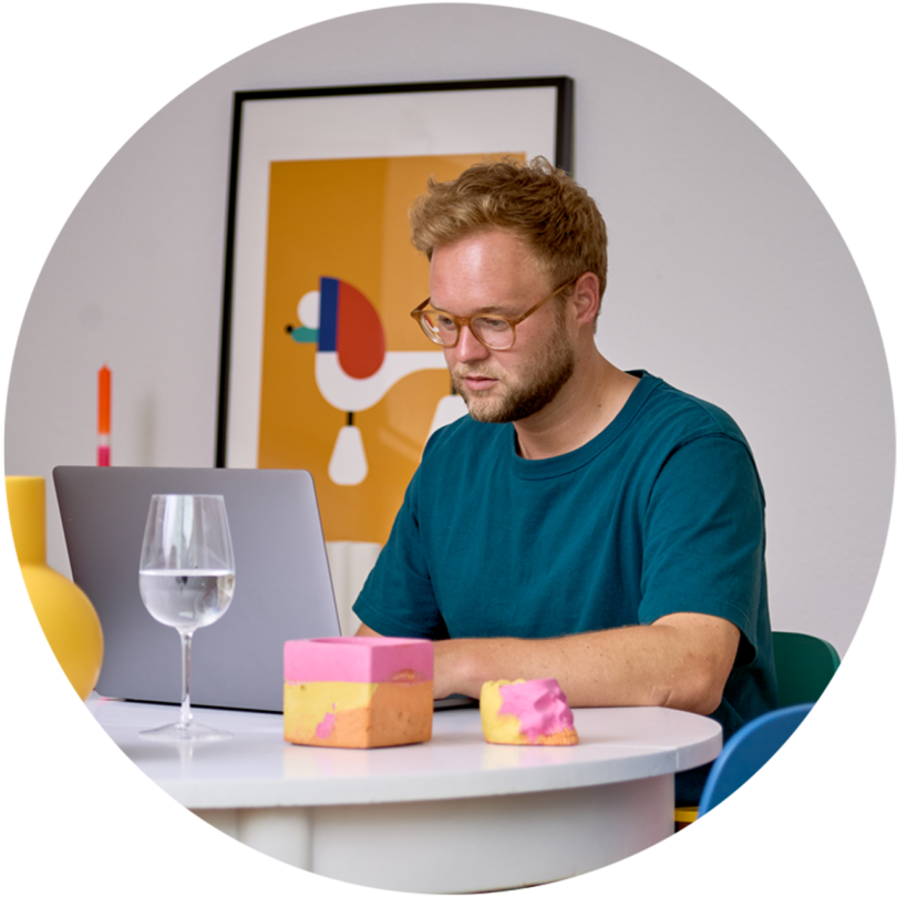 light-skinned male with light brown short hair wearing glasses and a turquoise t-shirt while sitting at a laptop on a table