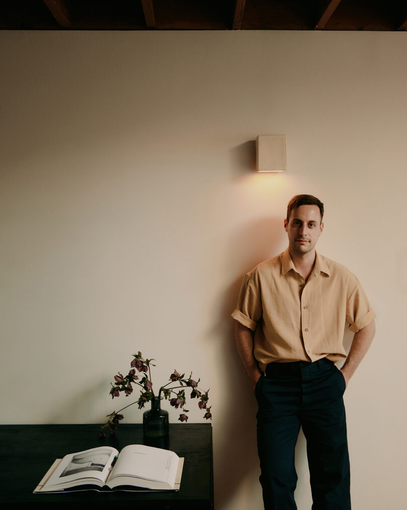 a light-skinned man with dark wearing a short-sleeved beige button-down short and black pants leans against a wall