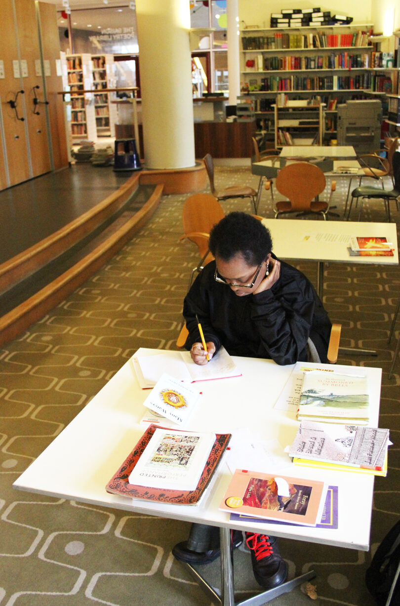 brown-skinned woman with short black hair and glasses sitting at a table covered in books and papers