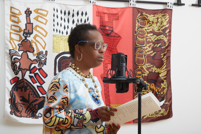 brown-skinned woman with short black hair and glasses stands in front of a wall of colorful tapestries reading to an audience