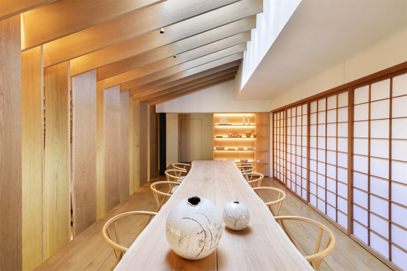 Dining room with long narrow wooden table with 8 chairs and round ceramic vases with shoji screen to the right. 