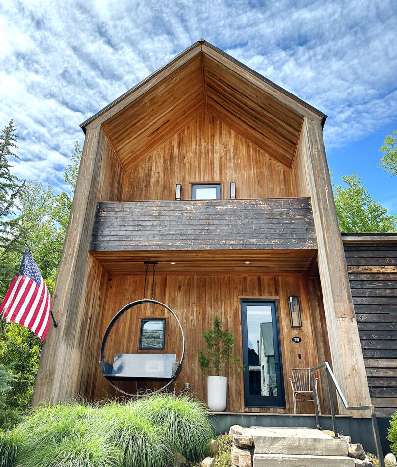 Wooden clad modernist home with American flag outside and large circular swinging bench out front.