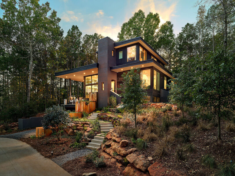 2021 Atlanta Homes & Lifestyles Serenbe Designer Showhouse, with illuminated interior and trees in the background with sunset skies glowing behind the home.