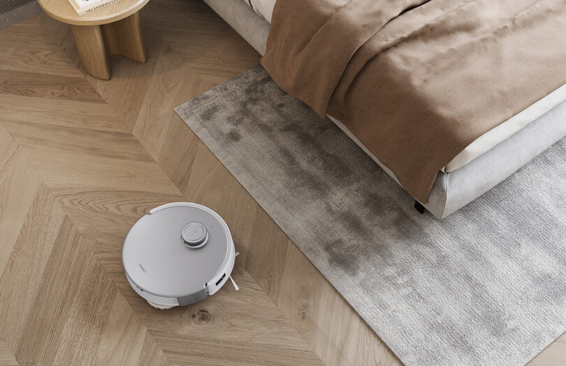 Robot vacuum cleaning hardwood floor and navigating toward gray low tuft rug underneath a bed.