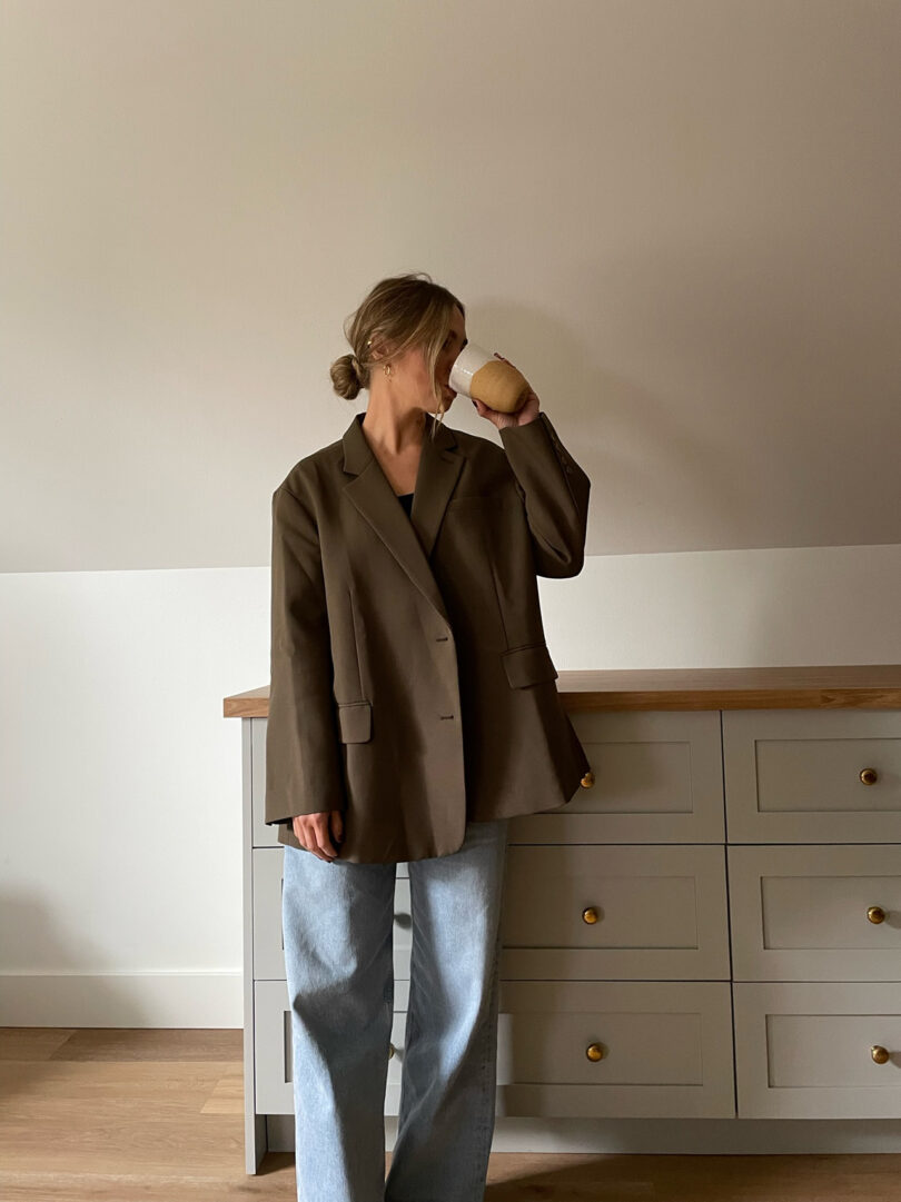 light-skinned woman wearing an oversized button-down shirt and jeans drinks from a cup of to-go coffee