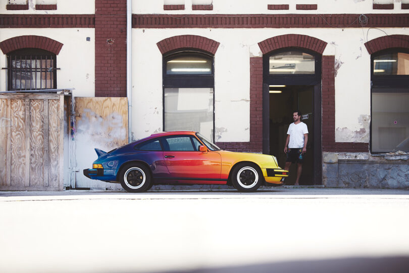 Porsche 911 painted in a gradient of color from blue to pink to yellow outside of a white and brown building