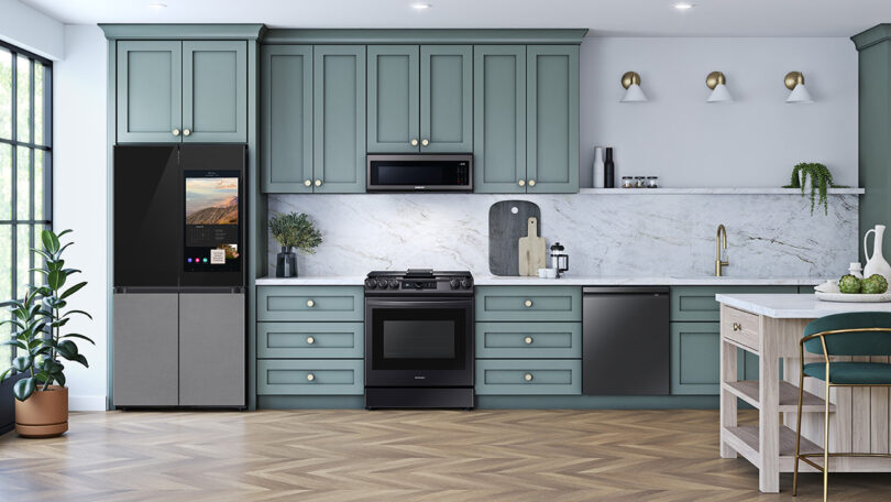 Large open contemporary kitchen with marble backsplash, herringbone pattern wood flooring, and counters and green-gray cabinetry, with Samsung Bespoke refrigerator inset into cabinetry to the left.