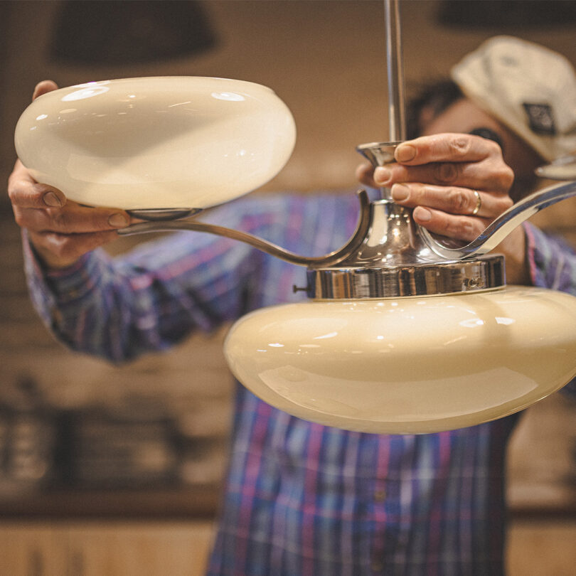 A white man in a shirt and a cap is adjusting a chandelier 